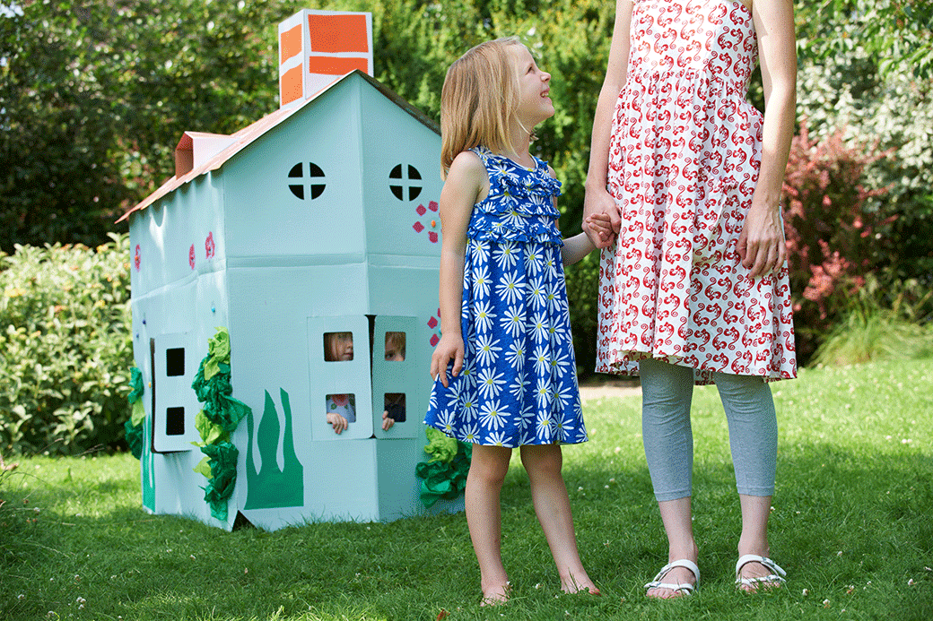 A little girl in a blue dress and an adult in a red dress hold hands in front of a white playhouse on a sunny day.